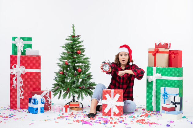 坐着前视图的年轻女子坐在圣诞礼物周围拿着白色墙上的时钟圣诞快乐时钟冬天