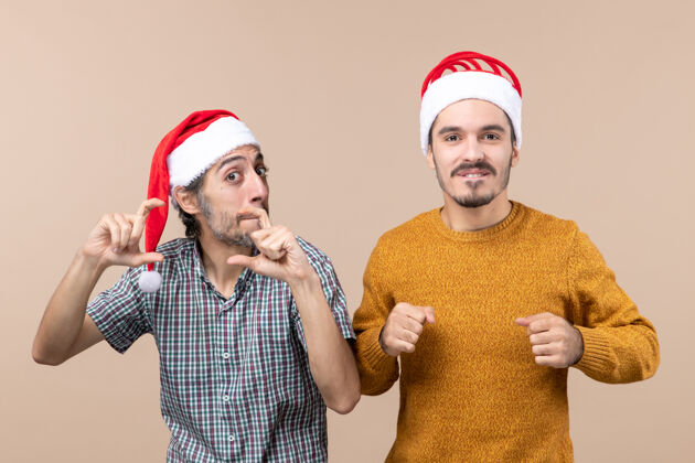 手正面图两个戴着圣诞帽的有趣的家伙 其中一个正在用手在米色孤立的背景上制作相机标志帽子两个风景