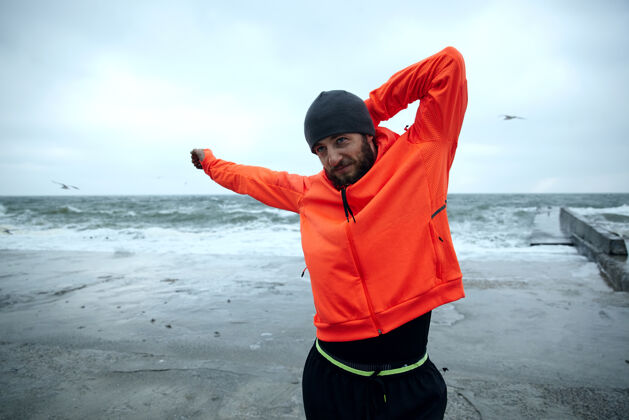 胡子年轻帅气的运动型黑发胡须男士在户外做伸展运动 准备晨练 身着黑色保暖运动服和带风帽的橙色外套男性暴风雨海边