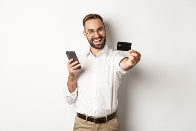 成人商务和在线支付兴奋的男人一边拿着智能手机一边出示信用卡 站着满意支付自信男人