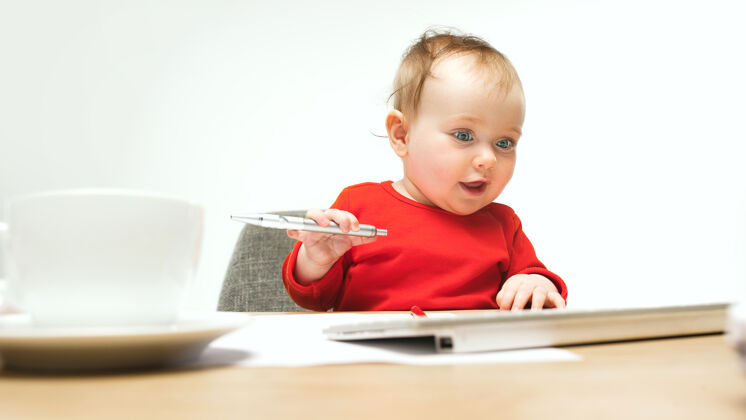 年轻快乐的小女婴坐在一个白色的工作室里 用现代电脑或笔记本电脑的笔和键盘开朗游戏电脑