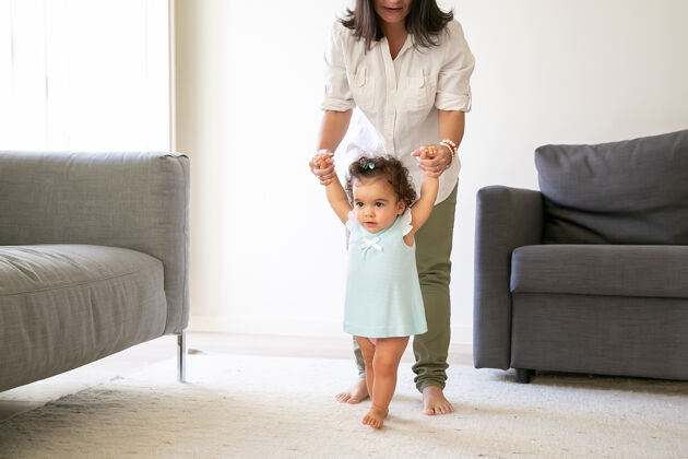 成长专注的女婴穿着淡蓝色的裙子 牵着妈妈的手 试图在家里散步全长亲子关系和童年的概念视图行走支持