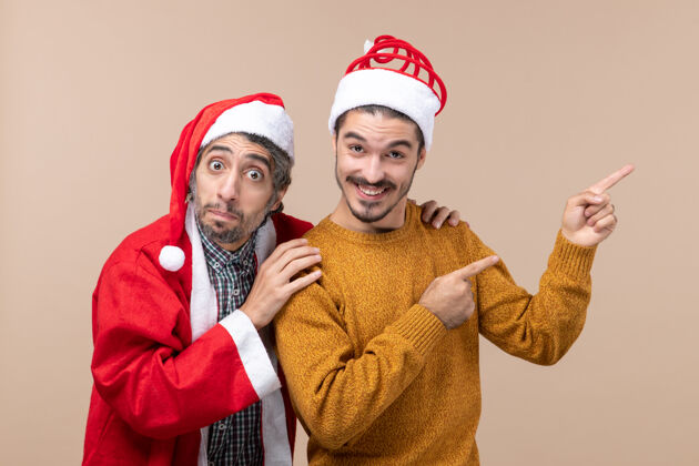 肩膀正面图：两个圣诞老人戴着圣诞帽 一个抱着朋友的肩膀 另一个在米色孤立的背景上显示方向帽子帽子前面