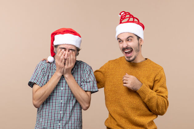 掩护前视图两个戴着圣诞帽的家伙一个用手捂住脸 另一个在米色孤立背景上大笑大笑男性视图