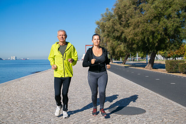 前面视图一对专注的成熟慢跑者沿着河岸奔跑白发男女穿着运动服 在户外跑步活动和退休概念前面女人灰色