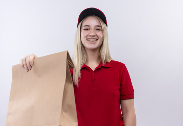 年轻微笑的小女孩穿着红色t恤 戴着帽子 戴着牙套 在隔离的白色背景下 站在相机的纸口袋里白色T恤拿着