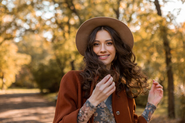 秋天画中有魅力的时尚微笑女子留着长长的卷发漫步在公园里穿着温暖的棕色外套秋日时尚 街头风格戴着帽子女孩服装装束