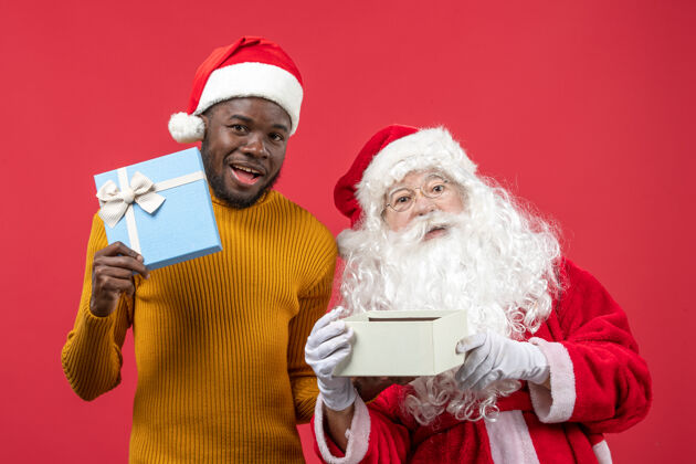 人红墙上的圣诞老人和拿着礼物的年轻人的正视图礼物前面肖像
