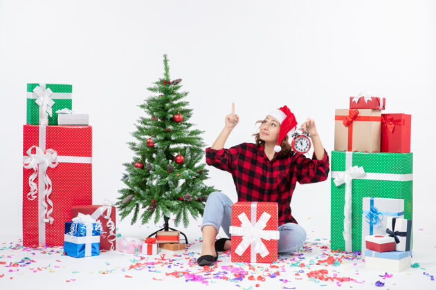 举行前视图的年轻女子坐在圣诞礼物周围拿着白色墙上的时钟礼物冬天圣诞树