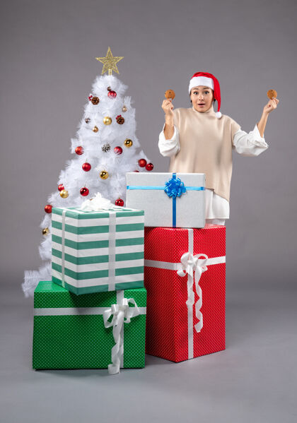 购物在圣诞树旁的灰色地板上 年轻的女人拿着饼干和礼物衣服包圣诞老人