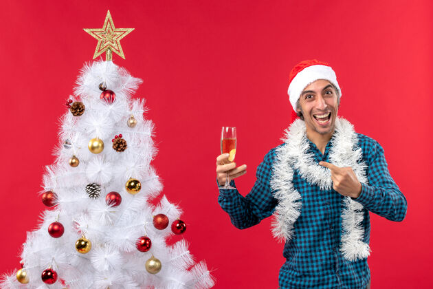 年圣诞心情快乐的年轻人 戴着圣诞老人帽 穿着蓝色条纹衬衫 在圣诞树旁拿着一杯葡萄酒年轻人人年