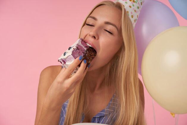 吹特写镜头：美丽的长发金发女人在享受美味的生日蛋糕 闭上眼睛 在粉色背景下展示她愉快的情绪 在五颜六色的气球里摆姿势女性饮食微笑