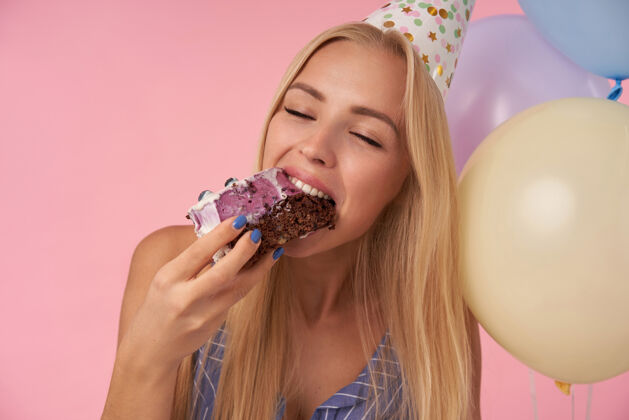饮食摄影棚拍摄了一位快乐的年轻长发女士 她闭着眼睛吃着节日蛋糕 在生日派对上度过了快乐的时光 站在粉色背景和氦气球上件庆祝气球