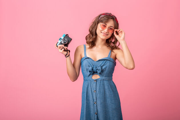 女人迷人的微笑女人在老式相机上拍照穿着牛仔裙和太阳镜隔离在粉红色的背景微笑开朗情感