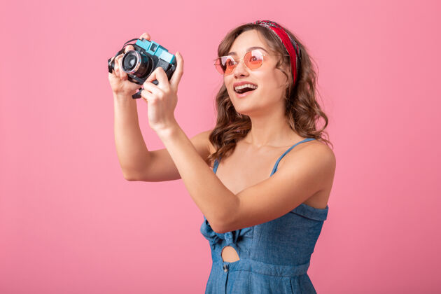 带着迷人的微笑女人在老式相机上拍照穿着牛仔裙和太阳镜隔离在粉红色的背景太阳镜女孩女人