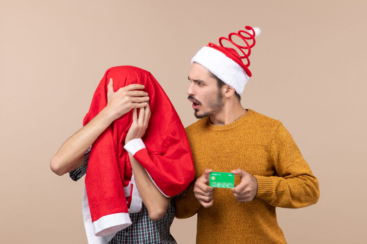圣诞老人正面图两个年轻人一个拿着信用卡 另一个用圣诞老人的外套盖住他的头 背景是米色的头前面肖像