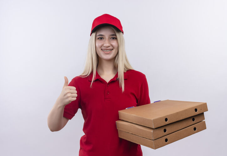戴着微笑着送货的年轻女孩穿着红色t恤 戴着帽子 戴着牙套 手里拿着披萨盒 在孤立的白色背景上竖起大拇指红色披萨女孩
