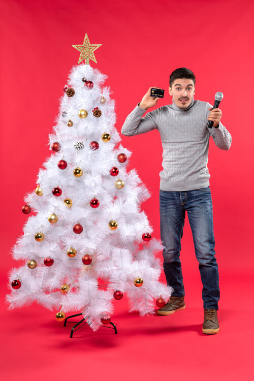 红色圣诞节心情自信的家伙穿着牛仔裤站在装饰圣诞树附近 拿着麦克风和手机微笑圣诞节心情