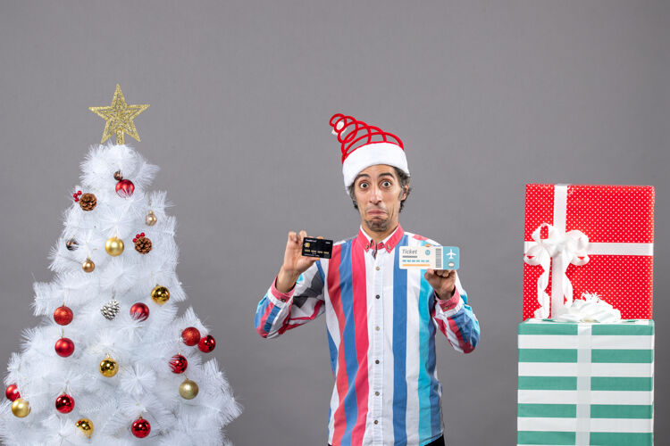 票前视图：一个眼睛敏锐的男人拿着卡片和旅行票 围着圣诞树和礼物人眼睛灰色