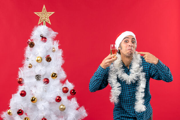 圣诞老人圣诞心情快乐疯狂情绪化的年轻人戴着圣诞老人帽 穿着蓝色条纹衬衫 举起一杯葡萄酒指着圣诞树快乐年新