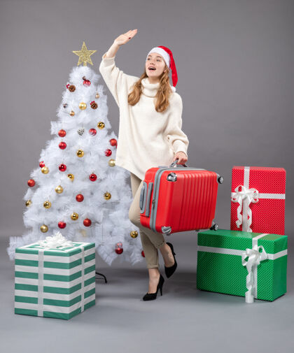 持有前视图欢呼的女孩与圣诞老人帽子举行她的旅行袋附近的白色圣诞树旅行礼物圣诞帽