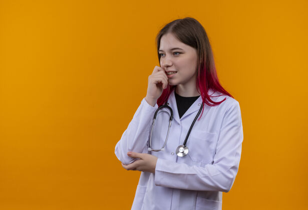 女孩看着身边年轻的医生女孩穿着听诊器医用长袍 手放在下巴上 背景是孤立的橙色 有复印空间听诊器下巴医学