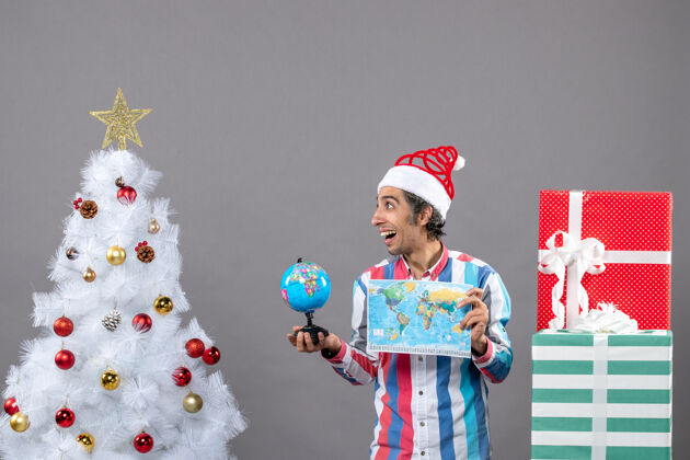 人前视图满意的男子与螺旋弹簧圣诞帽举行世界地图和地球仪圣诞树地图帽子