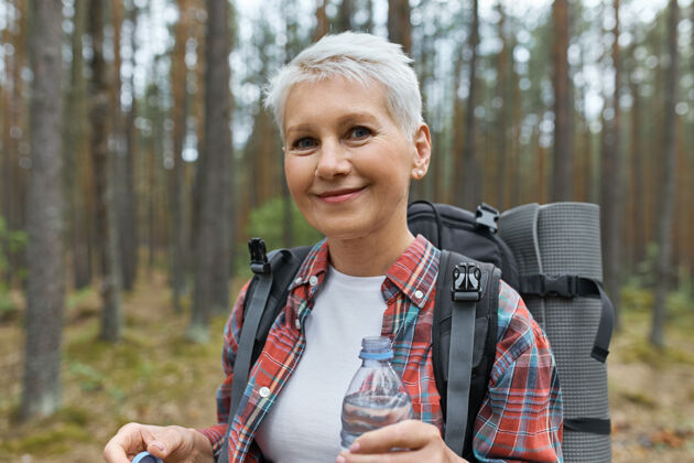人美丽的金发退休女性在户外度假 背着背包 拿着塑料瓶水在森林里散步旅游旅行者冒险