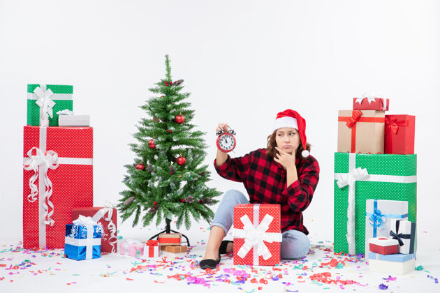 节日前视图的年轻女子坐在圣诞礼物周围拿着白色墙上的时钟庆祝十二月抱着