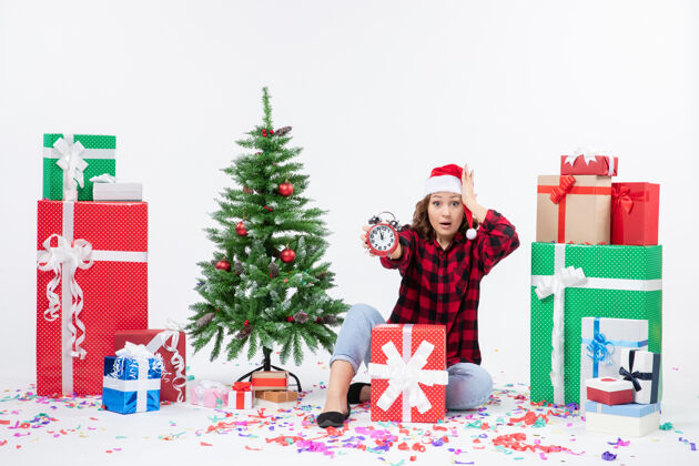 礼物前视图的年轻女子坐在圣诞礼物周围拿着白色墙上的时钟圣诞快乐抱着十二月