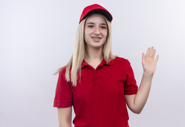 白色微笑送货的年轻女孩穿着红色t恤 戴着帽子 戴着牙套 在孤立的白色背景上显示问候手势手势微笑T恤