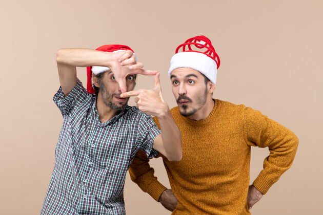 帽子正面图：两个戴着圣诞帽的好奇的家伙 一个用手在米色孤立的背景上制作相机标志帽子风景手