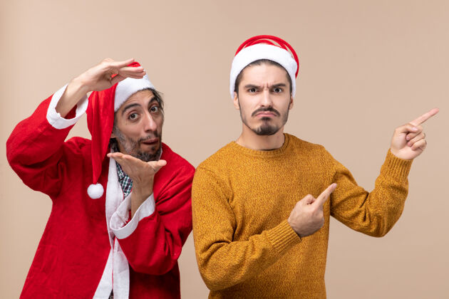 两个正面图两个戴着圣诞帽的朋友 一个表情严肃地在米色孤立的背景上展示方向男人米色圣诞老人