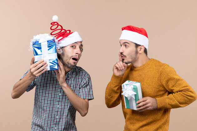 风景前视图两个困惑的家伙一个在检查他的圣诞礼物 另一个在米色背景上做嘘标志圣诞节礼物男人