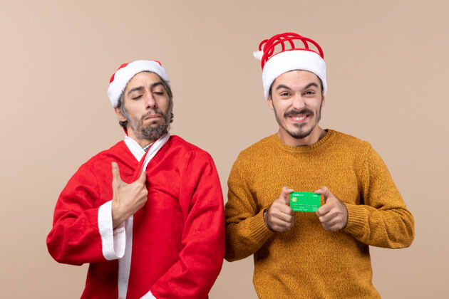 外套前视图两个朋友一个穿着圣诞老人的外套 另一个在米色孤立背景上的信用卡帽子信用卡米色