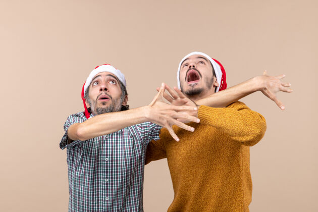 帽子正面图两个戴着圣诞帽的激动的家伙都躲着什么东西 同时看着高高的米色孤立背景高伙计们看