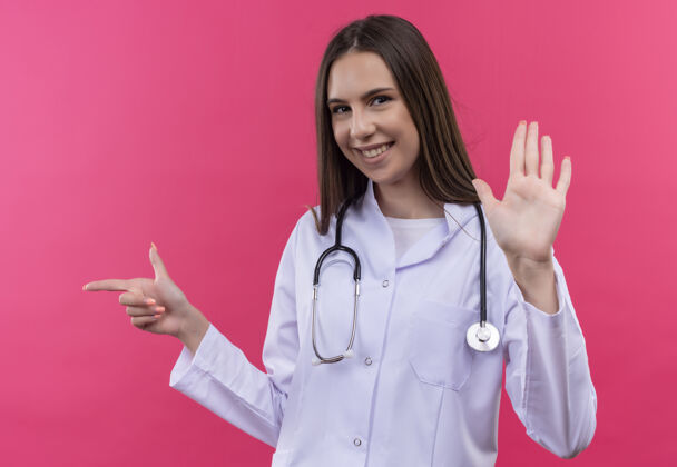 穿着微笑的年轻医生女孩穿着听诊器医用长袍指向侧面 在孤立的粉红色背景上显示停止手势听诊器侧面微笑