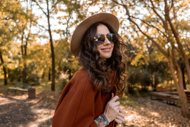 魅力迷人的时尚微笑的女人卷发漫步在街头公园穿着温暖的棕色外套秋季流行时尚 街头风格戴帽子和太阳镜波西米亚女士姿势