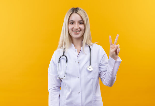 牙科微笑的医生穿着医用长袍戴着听诊器和牙套的年轻女孩在孤立的黄色背景上显示出和平的姿态和平女孩医生