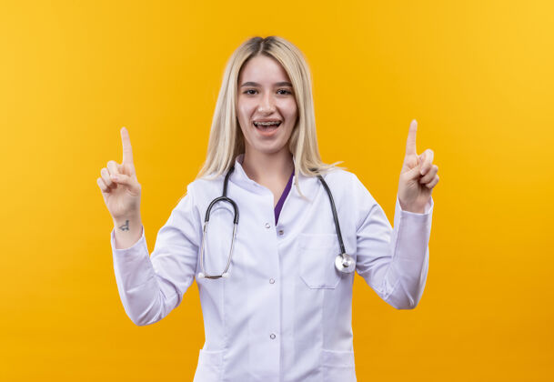 穿着微笑的医生穿着医用长袍戴着听诊器和牙套的年轻女孩指向孤立的黄色背景医生支架点