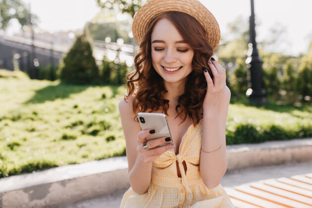 快乐快乐的红发女孩戴着可爱的帽子 带着快乐的微笑读着短信可爱的姜女穿着黄色的裙子在公园里放松的户外照片活动休息配件