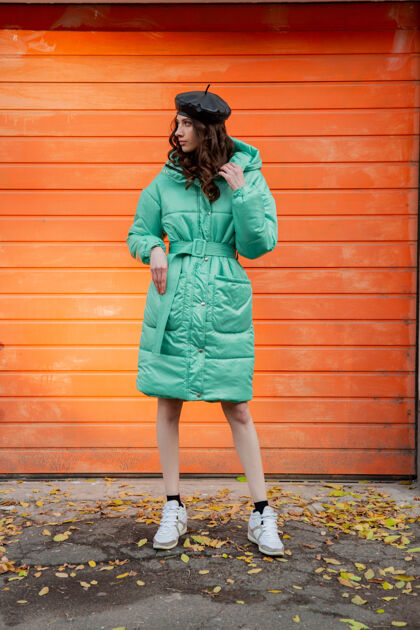 夹克穿着冬秋时尚潮流蓬松外套和帽子贝雷帽的时髦女人穿着运动鞋在街上对着橙色的墙壁摆姿势女人户外街头
