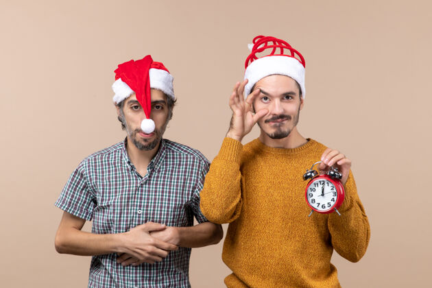 胸前前视图两个圣诞老人一个把手放在胸前 另一个拿着闹钟 背景是米色的手持帽子男人