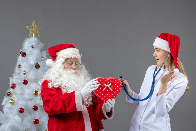 女圣诞老人和女医生的前视图 女医生在灰色墙上用听诊器观察礼物观察男人人们