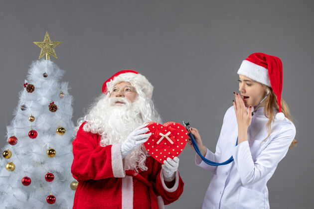 男性圣诞老人和女医生的前视图 女医生在灰色墙上用听诊器观察礼物男人圣诞老人女性