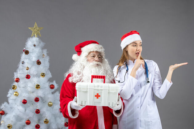 女圣诞老人和给他急救箱的女医生在灰色墙上的正视图谁人圣诞老人