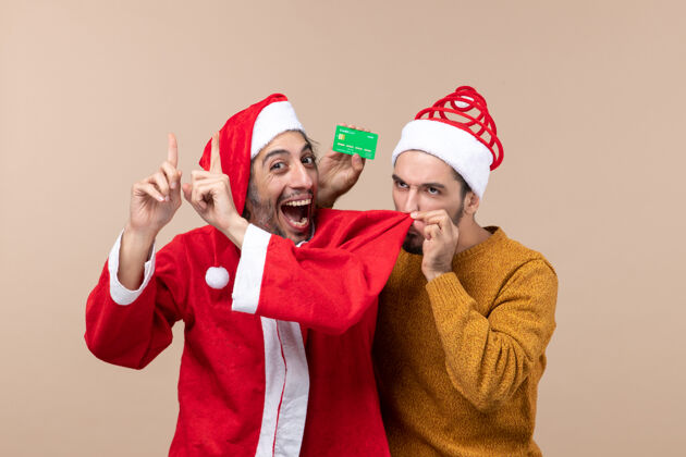 肖像前视图两个圣诞节的家伙一个穿着圣诞老人的外套 另一个用信用卡在米色的背景上嗅着他的朋友的外套成人圣诞节男性