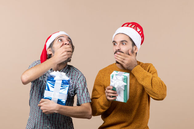 米色正面图两个戴着圣诞帽的震惊的家伙拿着圣诞礼物 手放在他们的嘴上 背景是米色的拿着圣诞老人帽子