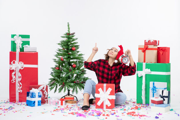 礼物前视图年轻女子坐在圣诞礼物周围拿着白色墙上的时钟节日快乐坐