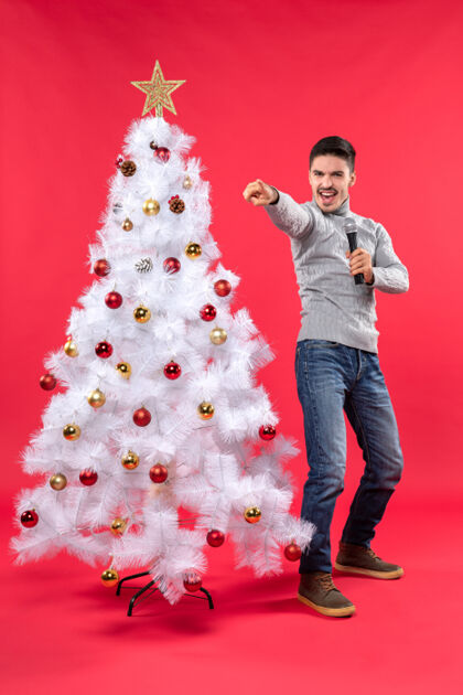 心情圣诞节的气氛与自信的家伙穿着牛仔裤站在装饰圣诞树附近 手持麦克风指向前方的东西衣服家伙庆祝